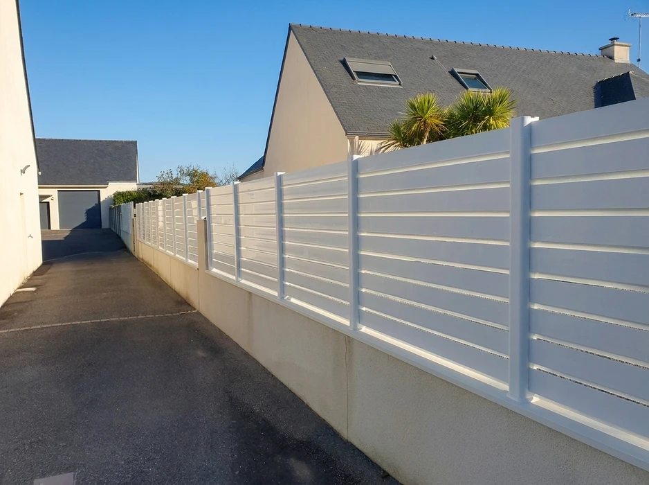 Clôture en aluminium blanc à lames horizontales, montée sur un muret en béton enduit blanc, longeant une allée bitumée entre deux maisons